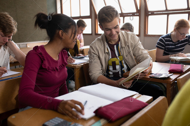 Side view of a teenage Caucasian boy and a teenage African American girl in a high school classroom sitting at desks, working together and reading, with teenage classmates sitting at desks working in the background