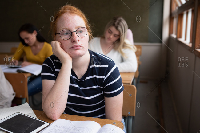 Side view of a teenage Caucasian girl in a school classroom sitting at desk, concentrating, with teenage male and female classmates sitting at desks working in the background
