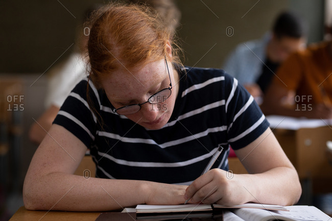 Front view close up of a teenage Caucasian girl in a school classroom sitting at desk, concentrating and writing, with teenage male and female classmates sitting at desks working in the background