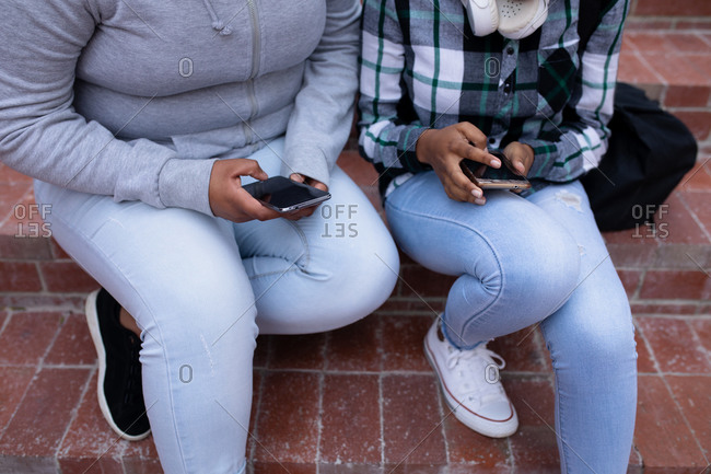 Low section front view close up of two African American teenage female high school pupils, using smartphones and sitting on steps in the school grounds