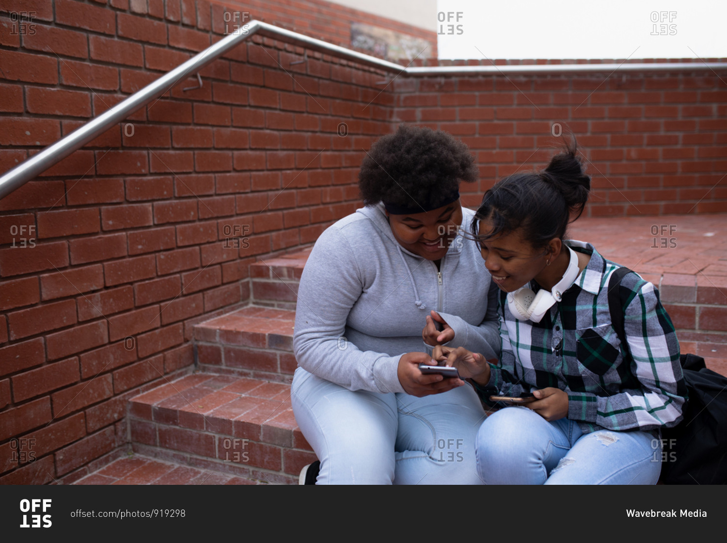 Side view of two high school African American teenage female school pupils, using a smartphone and sitting on steps in the school grounds