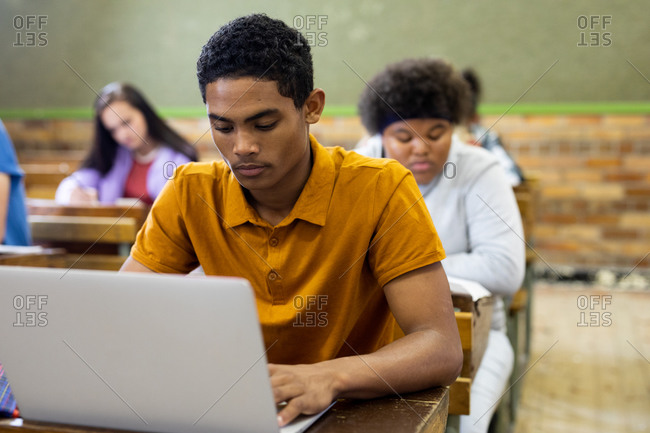 Front view of a teenage mixed-race boy in a school classroom sitting at desk, concentrating and using laptop computer, with teenage male and female classmates sitting at desks working in the background