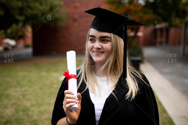 Front view of teenage Caucasian female high school student with long blonde  hair wearing a cap and gown, holding a diploma and smiling on her graduation  day stock photo - OFFSET