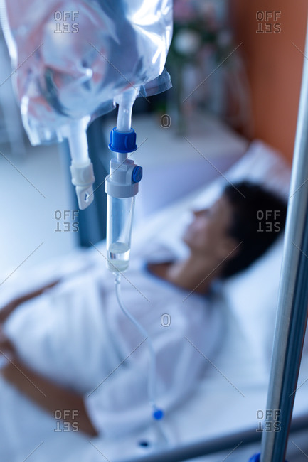 Close-up of intravenous drip with mixed-race female patient lying in bed on the background in the ward at hospital