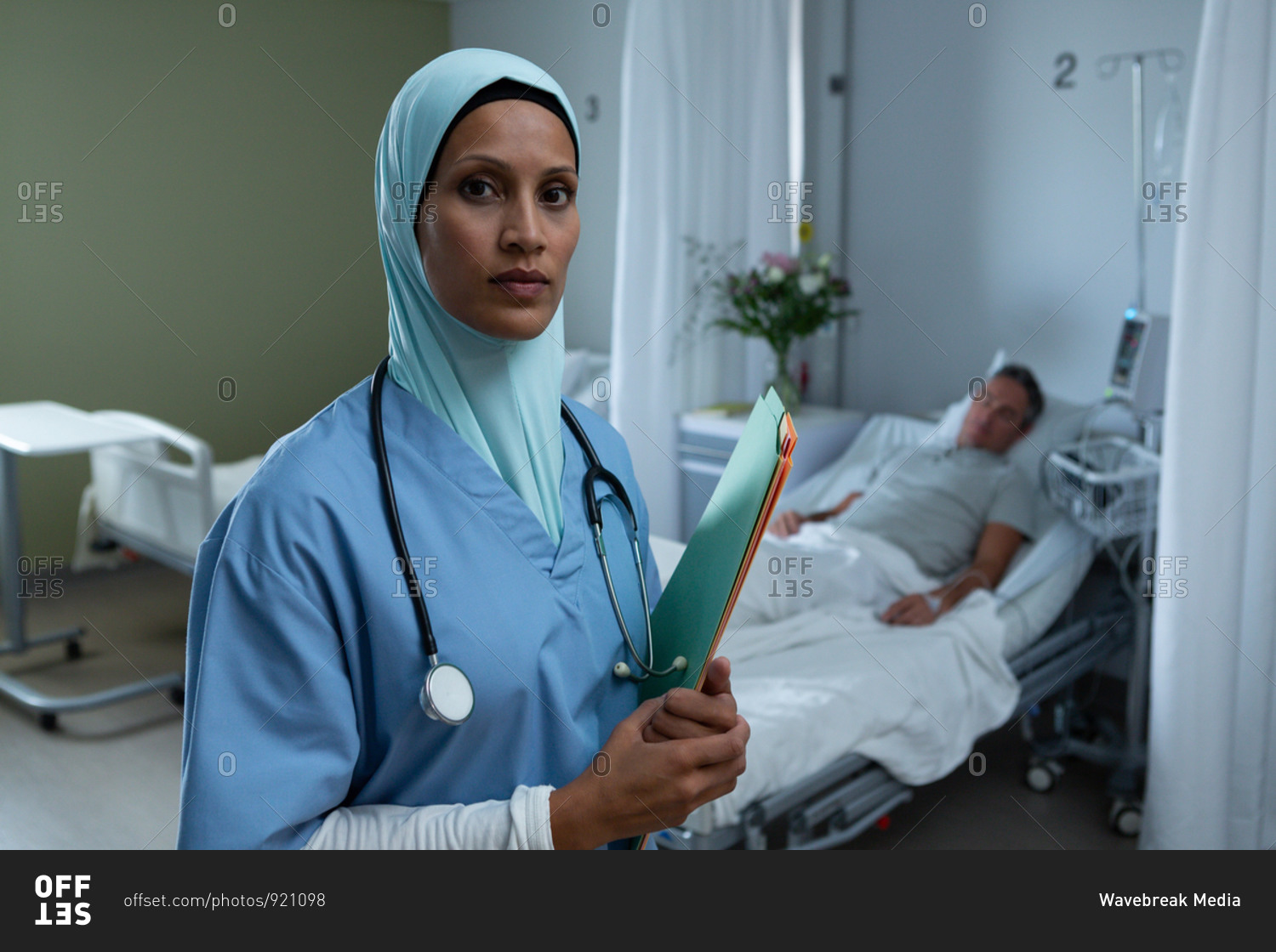 Portrait of beautiful mixed race female in hijab doctor standing with medical report while Caucasian male patient sleeps in the background in the hospital. Shot in real medical hospital with doctors nurses and surgeons in authentic setting