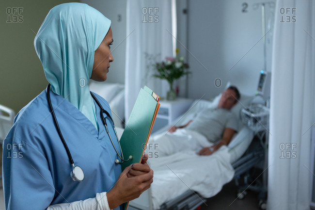 Side view of mixed race female doctor in hijab looking at Caucasian male patient who sleeps in bed in the ward in hospital. Shot in real medical hospital with doctors nurses and surgeons in authentic setting