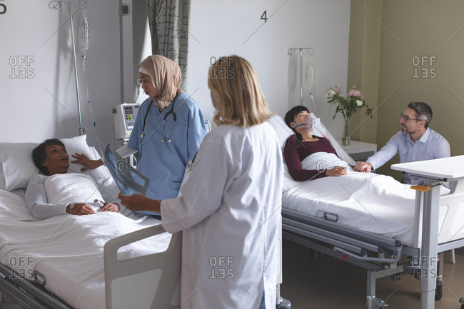 Side view of diverse female doctors interacting with female patient  in the ward at hospital. In the background Caucasian man is holding the hand of Asian woman who is lying in bed at hospital.