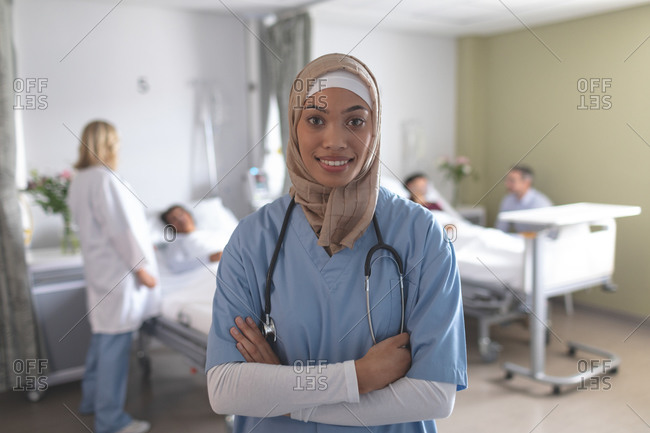 Portrait of mixed-race female doctor in hijab standing with arms crossed at hospital. In the background diverse doctors are interacting with their patients.