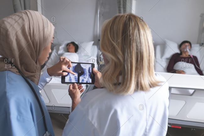 Rear view of diverse female doctors discussing over x-ray report on digital tablet in the ward at hospital. Diverse female patients are sleeping in bed in the background.