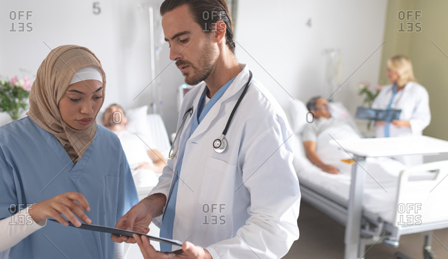 Side view of diverse doctors discussing over clipboard in the ward at hospital. In the background Caucasian female doctor discusses x-ray with senior mixed-race patient.