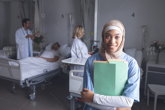Front view of mixed-race female doctor in hijab standing with medical files in the ward at hospital. In the background Caucasian doctors checking on patients.