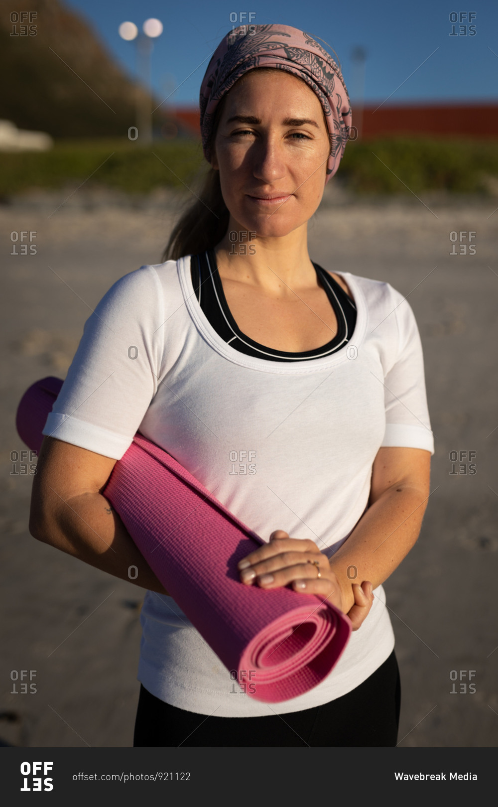 Portrait of a Caucasian woman, wearing white shirt and hair band, holding pink yoga mat under her arm, standing on a sunny beach.