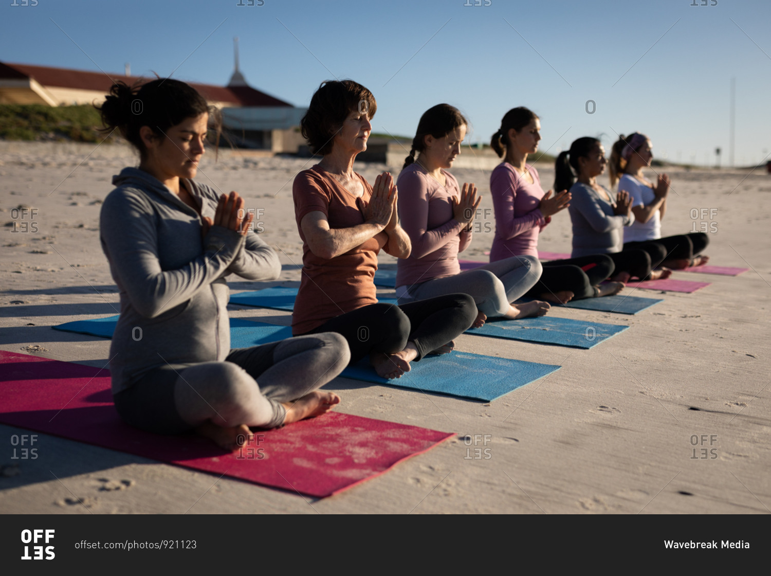 Side view of a multi-ethnic group of female friends enjoying exercising on a beach on a sunny day, practicing yoga sitting in a row in a yoga position, meditating with closed eyes and hands in prayer position.