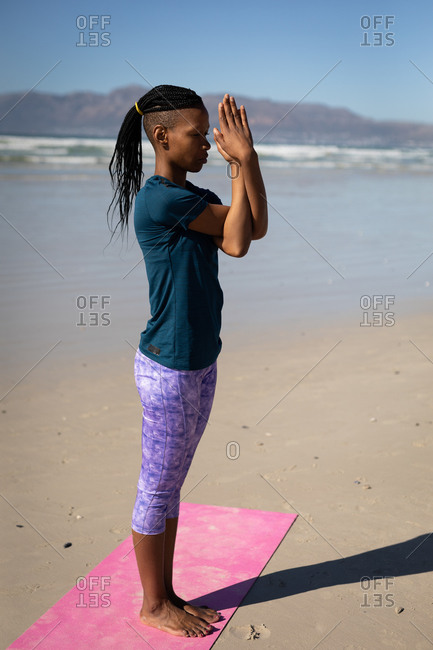 Side view of African American woman, wearing spots clothes, standing on pink yoga mat with arms in yoga position on the sunny beach.