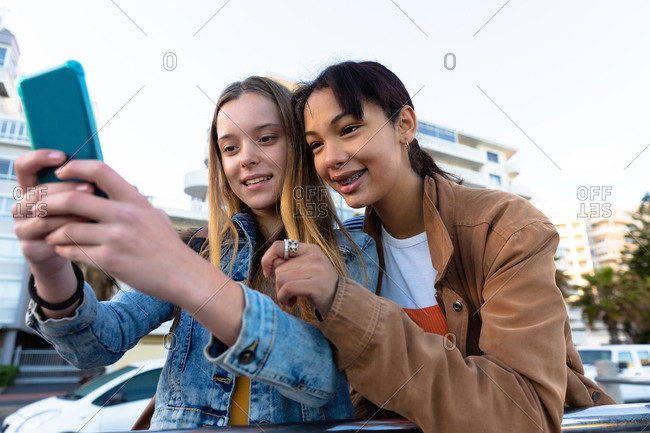 Front view of a Caucasian and a mixed race girl enjoying time hanging out together on a sunny day, standing and leaning on the railing, girl taking selfie of herself and her friend.
