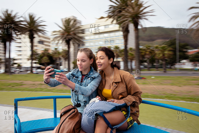Front view of a Caucasian and a mixed race girl enjoying time hanging out together on a sunny day on a playground, sitting on a merry-go-round, smiling, girl taking selfie of herself and her friend.