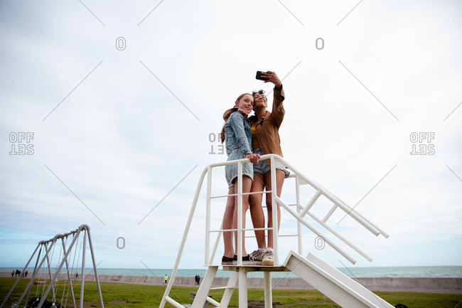 Front view of a Caucasian and a mixed race girl enjoying time hanging out together on a sunny day, standing together on the top on a slide, girl taking selfie of herself and her friend.