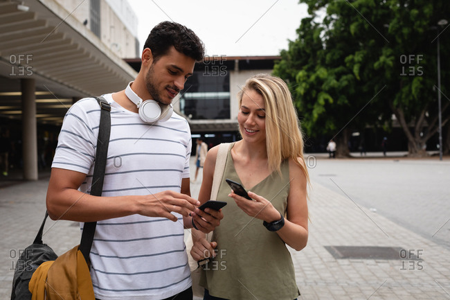 Front view of a Caucasian couple out and about in the city streets during the day, standing in the street and using their smartphones.