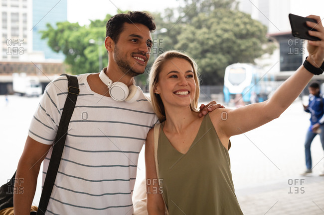 Front view of a happy Caucasian couple out and about in the city streets during the day, standing in the street and taking a selfie with their smartphone.