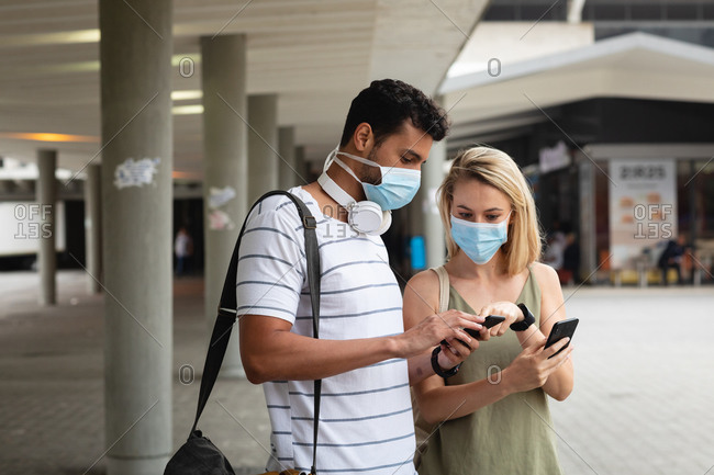 Front view of a caucasian couple out and about in the city streets during the day, wearing face masks against air pollution and covid19 coronavirus, using their smartphones.