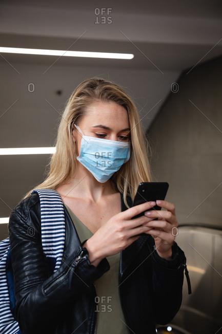 Front view of a caucasian woman with long blind hair, leaving an escalator, using her smartphone and wearing face mask against air pollution and covid19 coronavirus.
