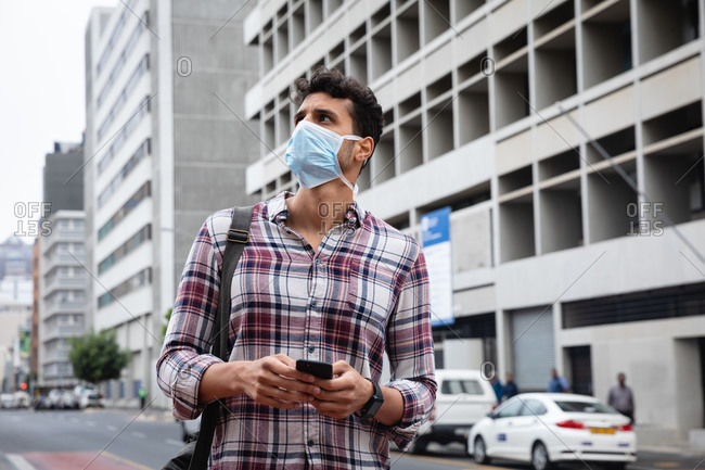 Front view close up of a caucasian man wearing checkered shirt and face mask against air pollution and covid19 coronavirus, walking through the city streets, using his smartphone.