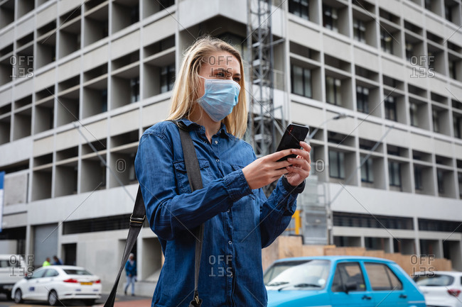 Front view close up of a caucasian woman wearing face mask against air pollution and covid19 coronavirus, walking through the city streets, using her smartphone.