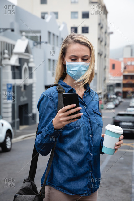Front view close up of a caucasian woman wearing face mask against air pollution and covid19 coronavirus, walking through the city streets, using her smartphone and holding a cup of takeaway coffee.