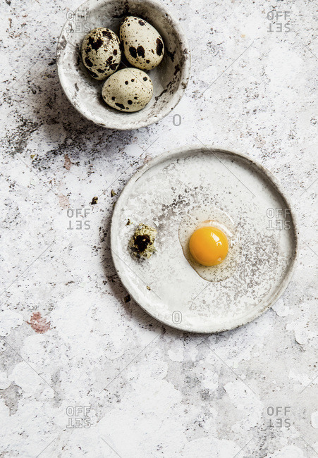 Cracked quail egg on a grey ceramic plates, few eggs in a small grey bowl, grey background.