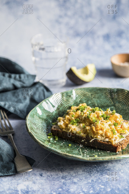 Breakfast. Scrambled eggs on rye bread with chive with avocado in the background and glass of water.