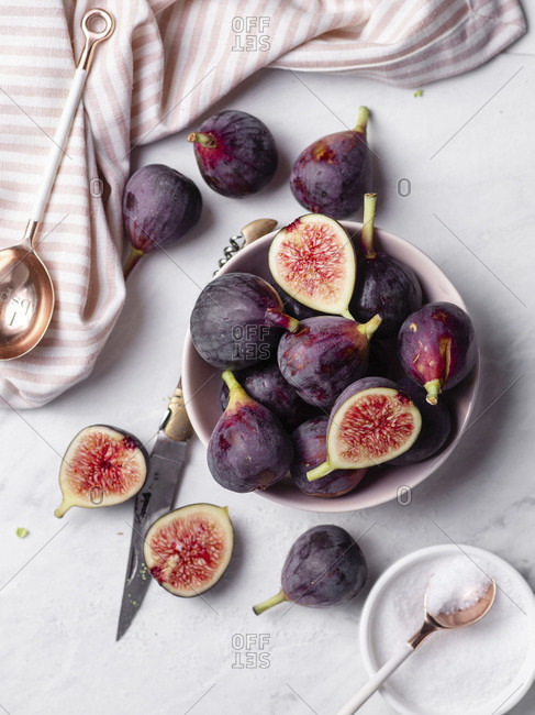 A bowl of ripe, purple figs on a white marble countertop surrounded by a striped napkin, rose gold spoons, and a small bowl of sugar