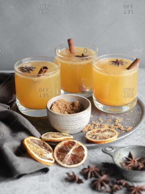 Three glasses of an orange spiced drink on a grey background, garnished with cinnamon sticks, brown sugar, blood oranges, and star anise.