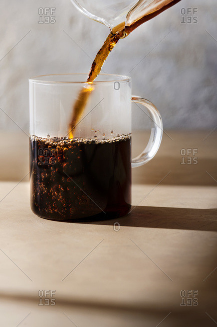 Hot coffee being poured into a glass mug. Light brown concrete surface with morning light coming through a window.