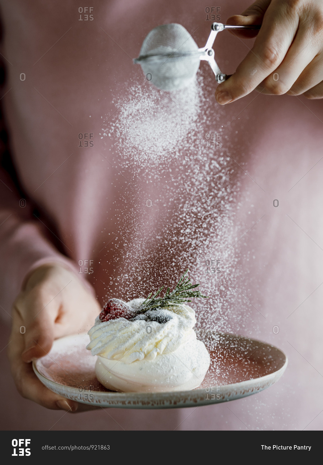Woman in pink blouse holds with one hand pink plate with mini Pavlova cake decorated fresh berries and rosemary, and sprinkles with her other hand icing sugar through a sieve.