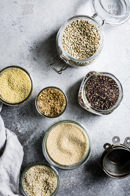 A collection of seeds and grains on a grey background, buckwheat, brown rice, quinoa, millet, amaranth, and flax seeds