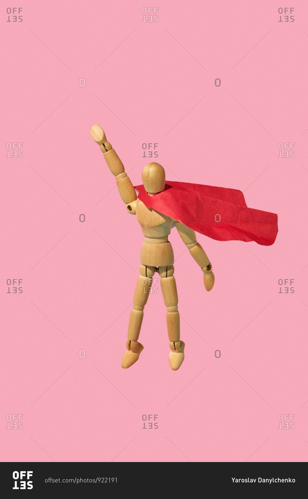 Movable miniature articulation mannequin in a red cape is flying up with raised hand as a superhero or superman against pink background.