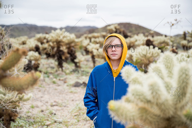 Tween boy with glasses behind cholla cactus in desert in front of hill