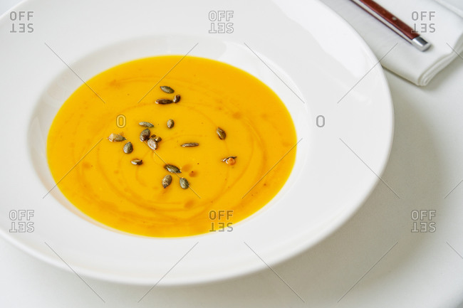Homemade creamy pumpkin soup topped with olive oil and toasted pumpkin seeds on white table