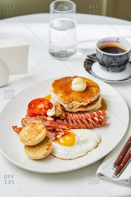 Full American Breakfast dish with a stack of pancakes, sausages, tomatoes, champignons, bacon and egg on white round table