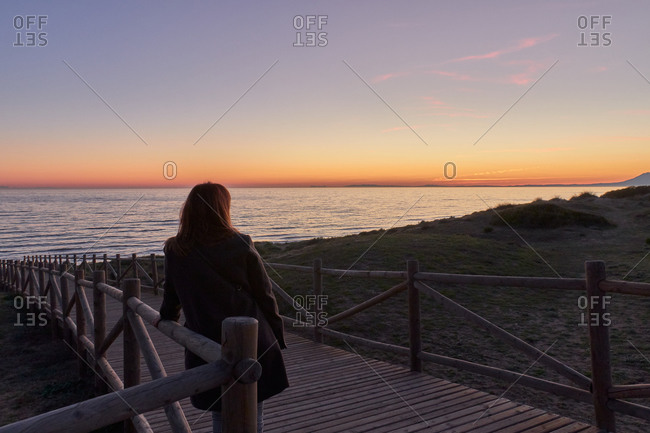 Woman looking sunset on boardwalk path leading to a beach with calm sea
