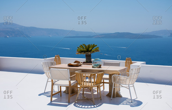 Furniture composed by a table and some chairs on a white terrace of a house in Santorini Greece where you can enjoy meal while seeing a romantic seascape to the blue Aegean sea. Horizontal ph