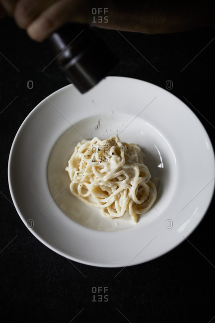 Sprinkling pepper onto a bowl of cacio e pepe pasta on dark surface