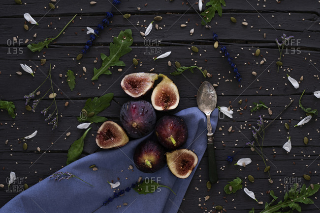 From above fresh halved figs and silver spoon placed on wooden table near blue napkin and arugula in kitchen