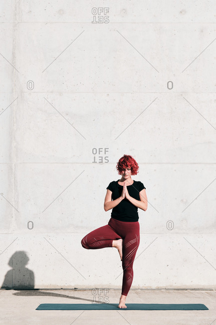 Confident barefoot female athlete in sportswear meditating while standing in rakshasa position with hands close to the chest in namaste on sports mat in front of concrete wall in sunlight