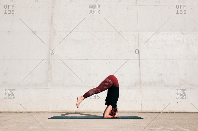 Side view of unrecognizable barefooted female athlete in activewear standing upside down in sirsasana position with legs raised on sports mat training alone on street against concrete wall in daytime