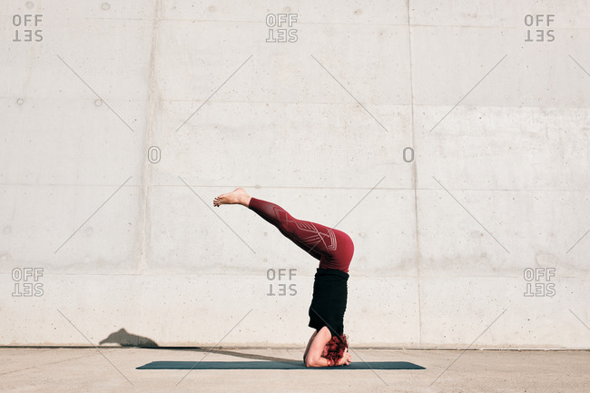 Side view of unrecognizable barefooted female athlete in activewear standing upside down in sirsasana position with legs raised on sports mat training alone on street against concrete wall in daytime
