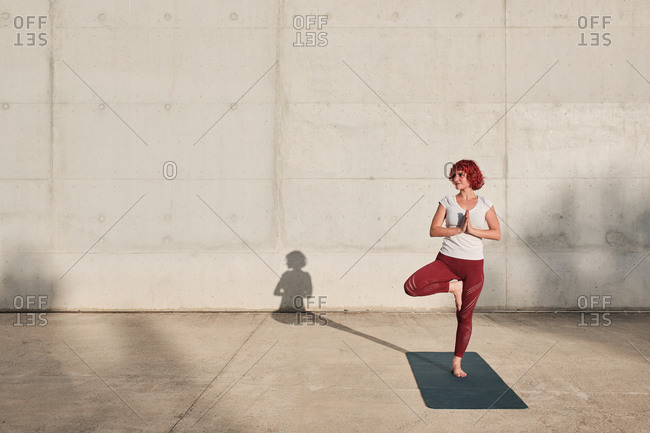 Barefoot female athlete in sportswear meditating while standing in rakshasa position with hands close to the chest in namaste on sports mat in front of concrete wall in sunlight looking away