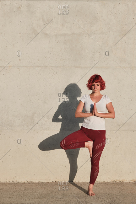 Peaceful barefoot female athlete in sportswear meditating while standing in rakshasa position with hands close to the chest in namaste on sports mat in front of concrete wall in sunlight