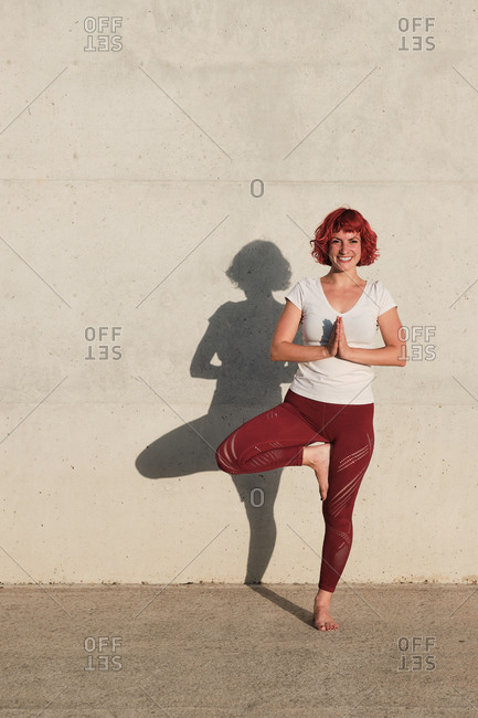Happy barefoot female athlete in sportswear meditating while standing in rakshasa position with hands close to the chest in namaste on sports mat in front of concrete wall in sunlight