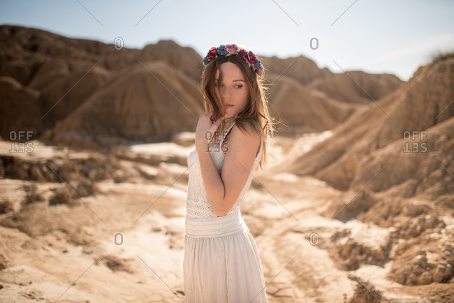 Side view of young woman in white dress and floral wreath touching hair and looking away while standing against rocky hills on sunny day in Bardenas Reales in Navarre, Spain